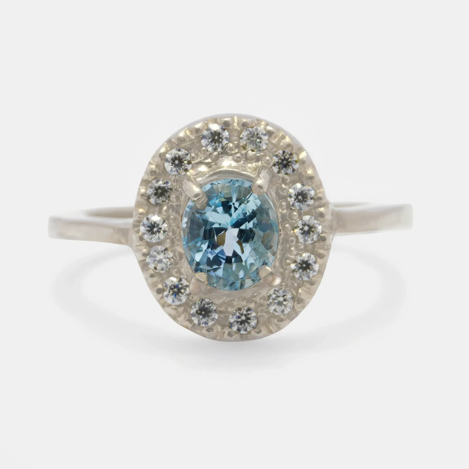 Anillo de plata visto de frente, con una piedra ovalada de color celeste sujeta por garras y un halo de piedras blancas alrededor.